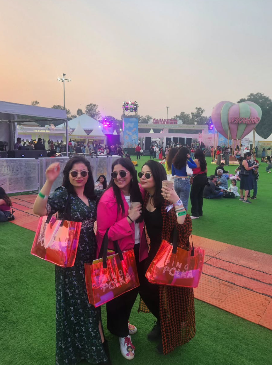 Three women posing with colorful bags at an outdoor event with a hot air balloon in the background.
